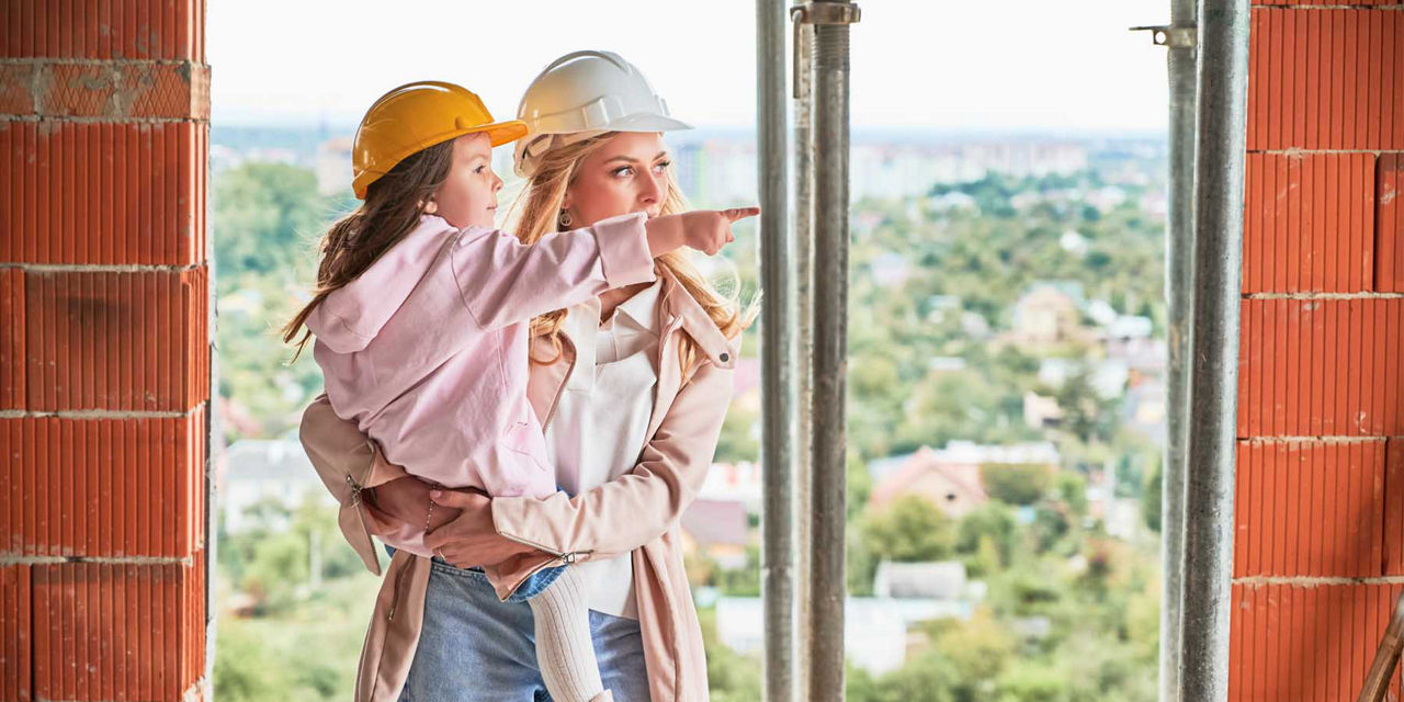 Junge Mutter hat ihre Tochter auf dem Arm und prüft den Hausbau-Fortschritt