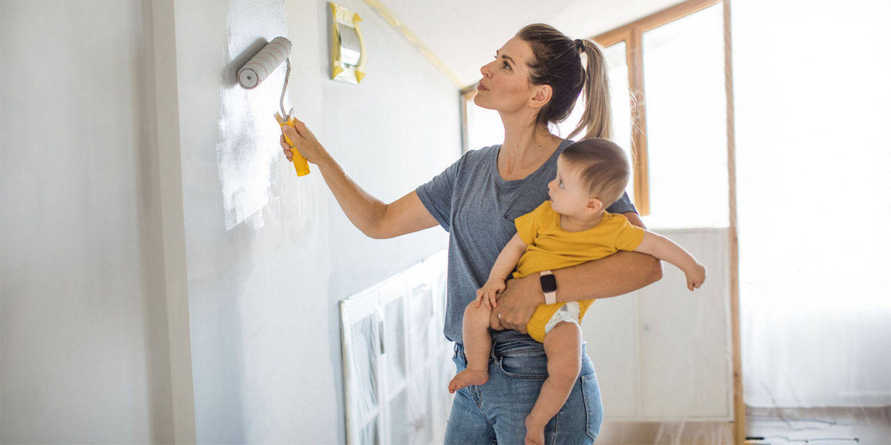 Mutter hat ihr Kind im Arm während sie eine Wand streicht