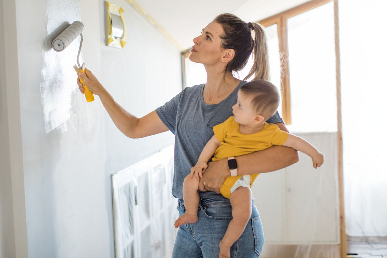 Mutter hat ihr Kind im Arm während sie eine Wand streicht