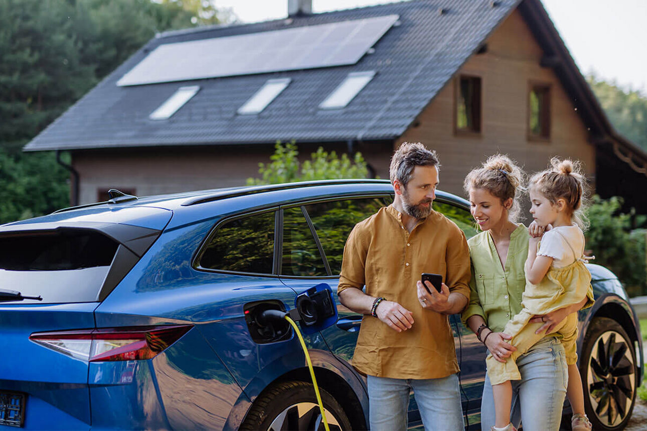 Junge Familie steht vor ihrem Einfamilienhaus mit Solaranlage und Elektroauto
