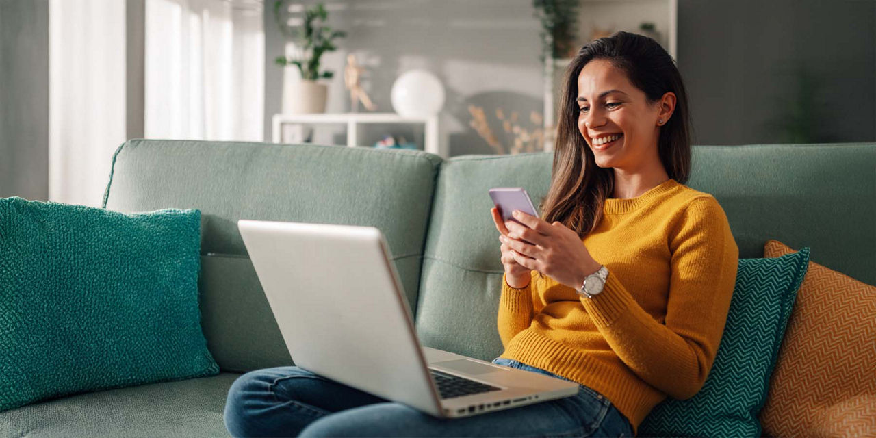 Junge Frau sitzt lächelnd auf ihrem Sofa und schaut auf ihren Laptop