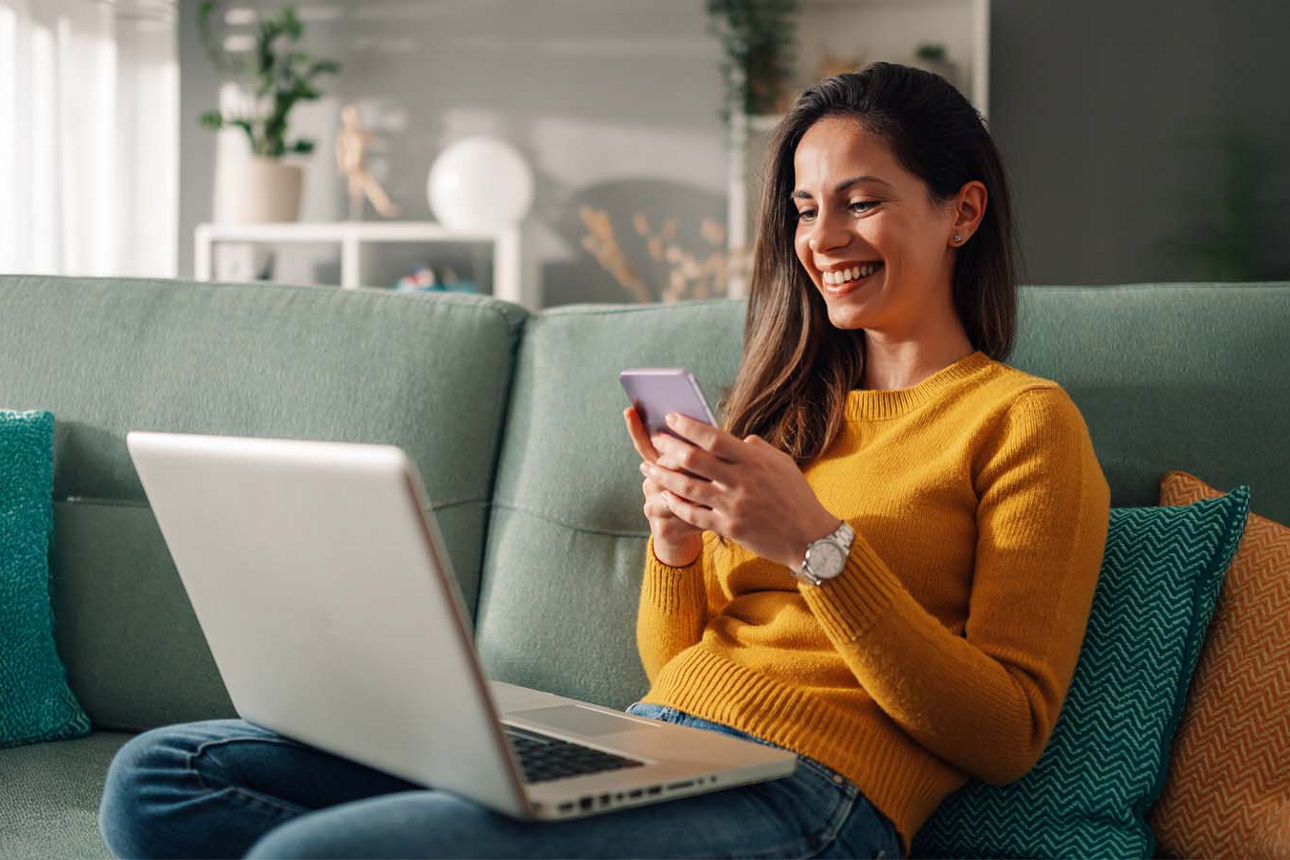 Junge Frau sitzt lächelnd auf ihrem Sofa und schaut auf ihren Laptop