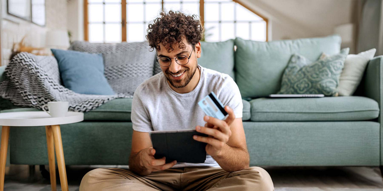Junger Mann mit Kreditkarte in der Hand schaut zufrieden auf sein Tablet