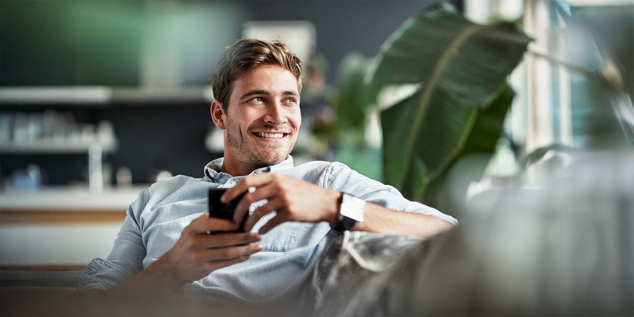 Mann sitzt lächelnd auf dem Sofa und hat sein Smartphone in der Hand