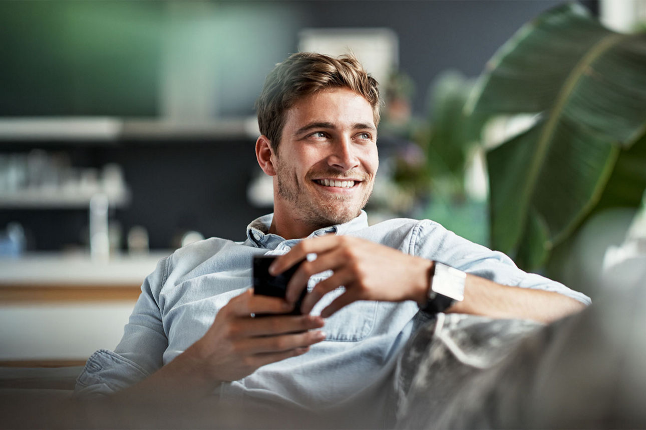 Mann sitzt lächelnd auf dem Sofa und hat sein Smartphone in der Hand