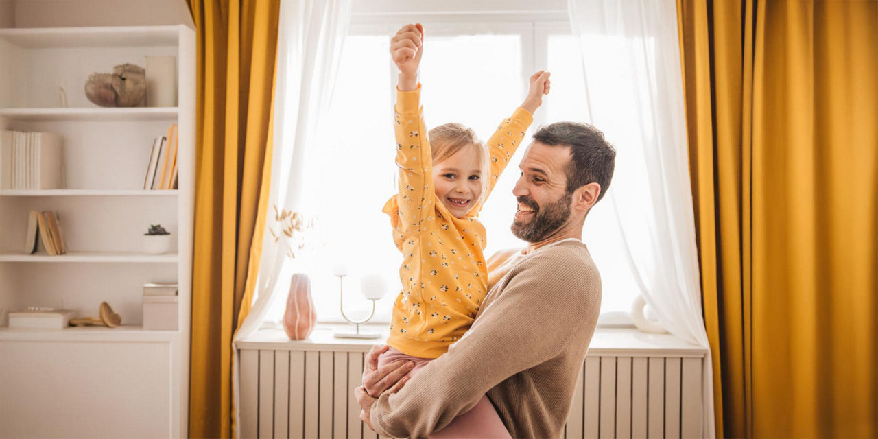 Ein Vater trägt lachend seine Tochter im Arm während diese die Arme in die Höhe reißt