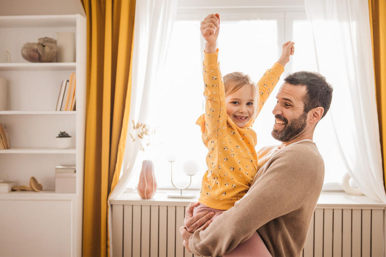 Ein Vater trägt lachend seine Tochter im Arm während diese die Arme in die Höhe reißt