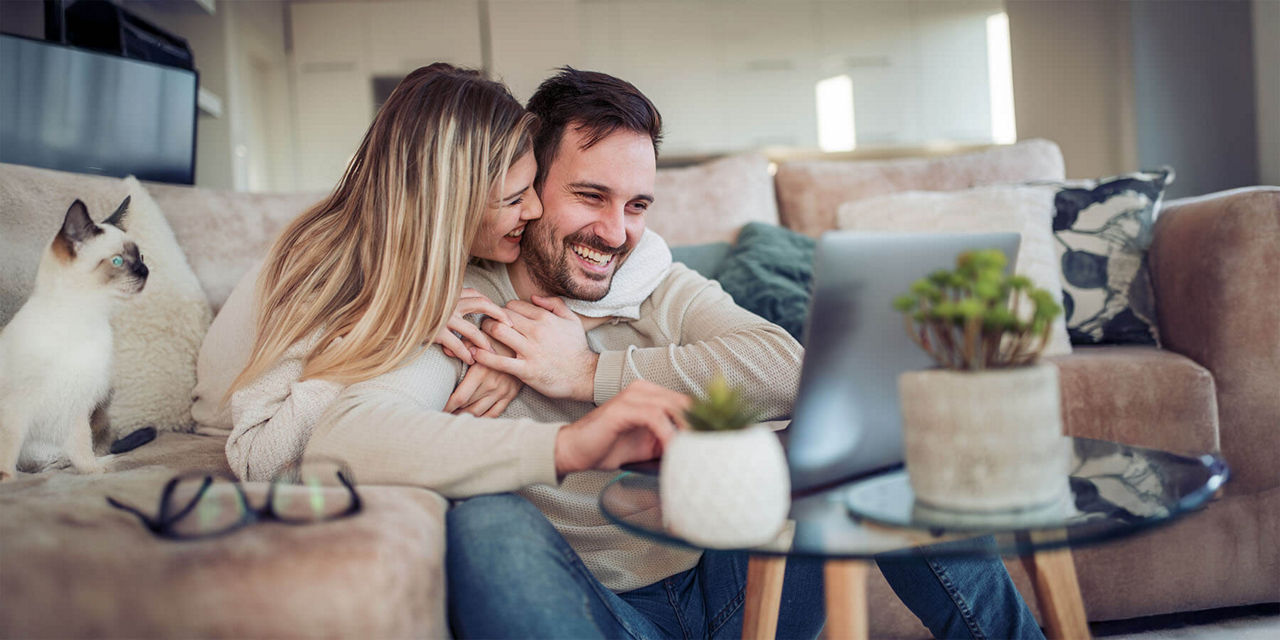 Junges Paar sitzt lachend auf dem Sofa und schaut auf den Laptop