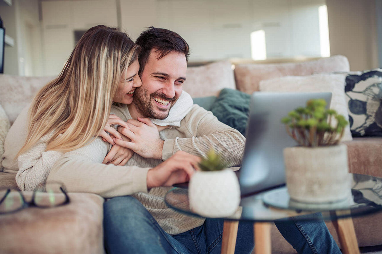 Junges Paar sitzt lachend auf dem Sofa und schaut auf den Laptop