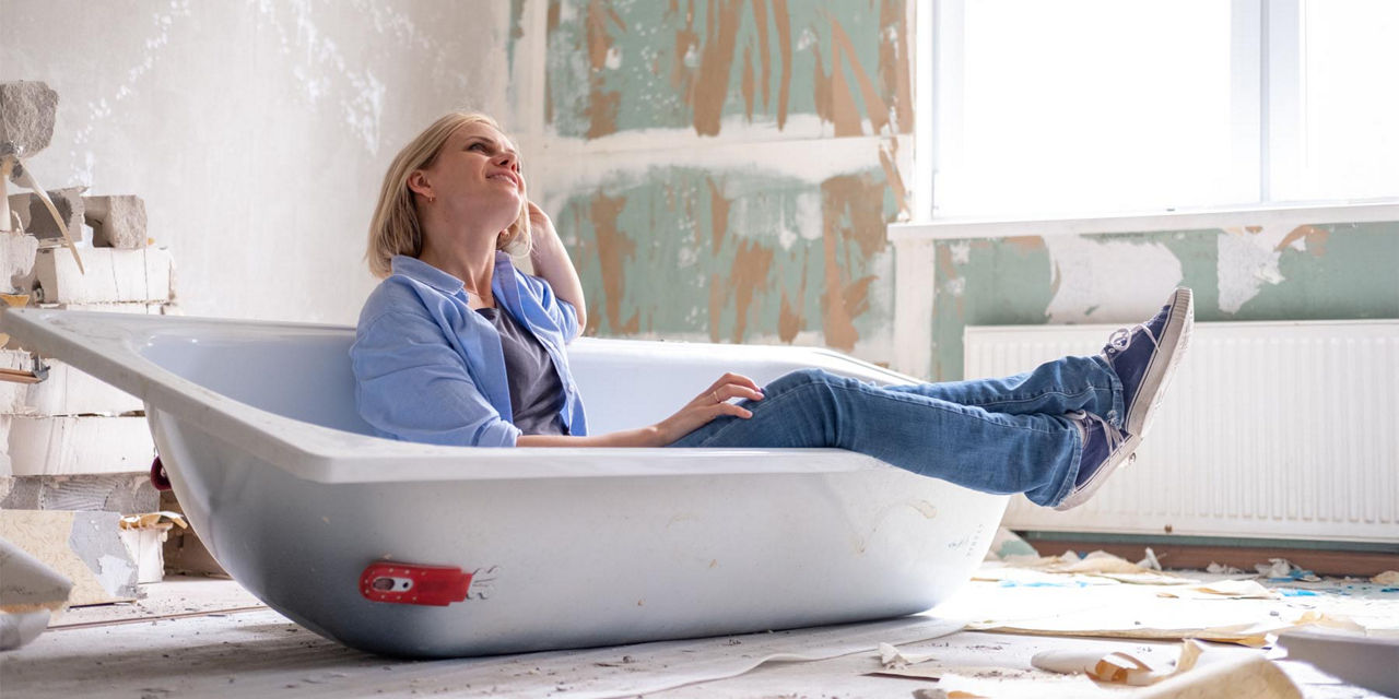 Junge Frau sitzt lächelnd in der Badewanne auf der Baustelle ihres Eigenheims