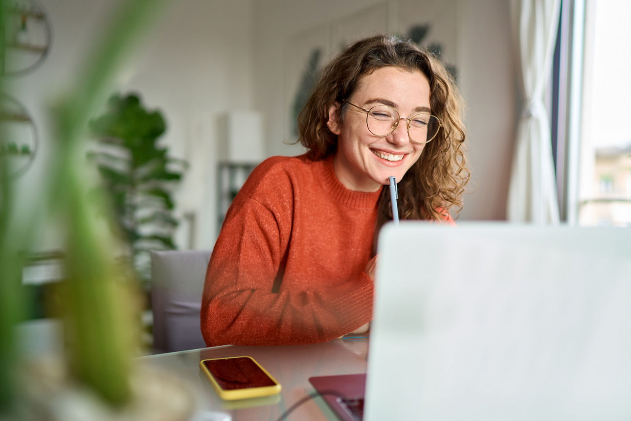 Eine junge Frau sitzt vor ihrem Laptop, hält einen Stift an ihr Kinn und lacht.