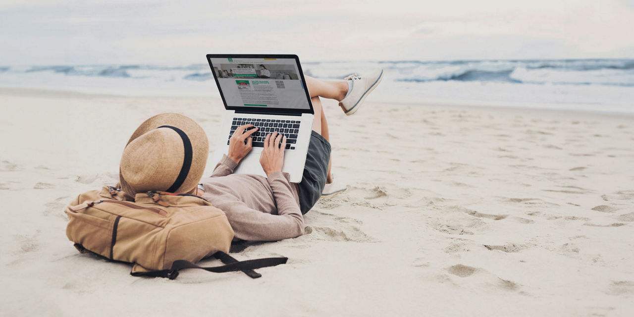 Person liegt in Sommerklamotten und Strohhut am Strand und arbeitet am Laptop