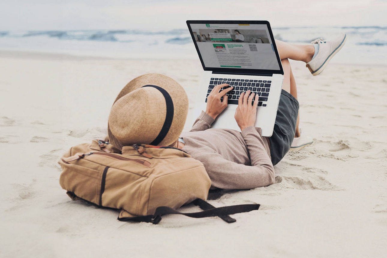 Person liegt in Sommerklamotten und Strohhut am Strand und arbeitet am Laptop