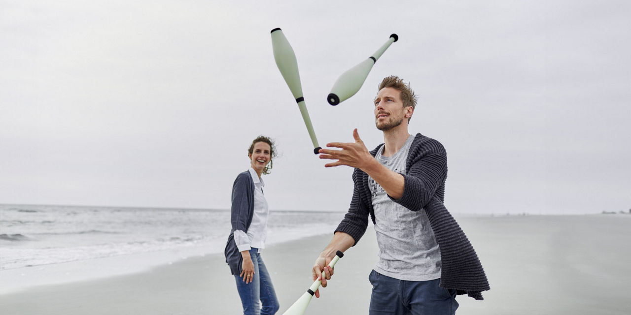 Mann jongliert am Strand und eine Frau schaut ihm dabei begeistert zu