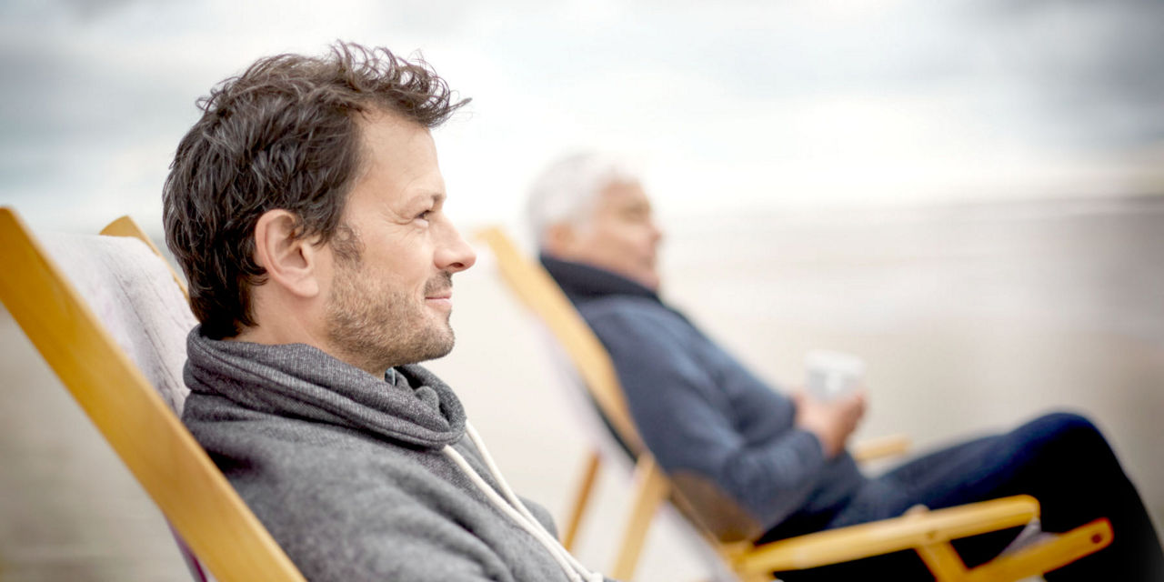 Vater und Sohn sitzen am Strand im Liegestuhl und trinken eine Tasse Kaffee