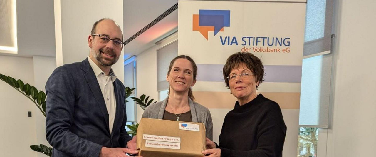 Ein Paket wird an das Frauenhaus in Warendorf übergeben. Links steht ein Mann, rechts daneben zwei Frauen. Sie halten das Paket gemeinsam in der Hand.