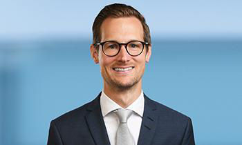Er hat braune Haare, trägt Brille und Anzug mit Krawatte