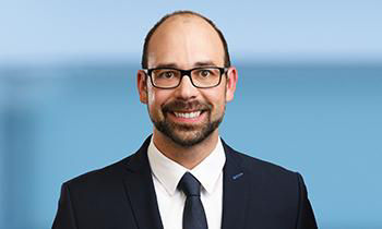 Er hat braune Haare, trägt Brille und Anzug mit Krawatte