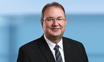 Er hat braune Haare, trägt Brille und Anzug mit Krawatte