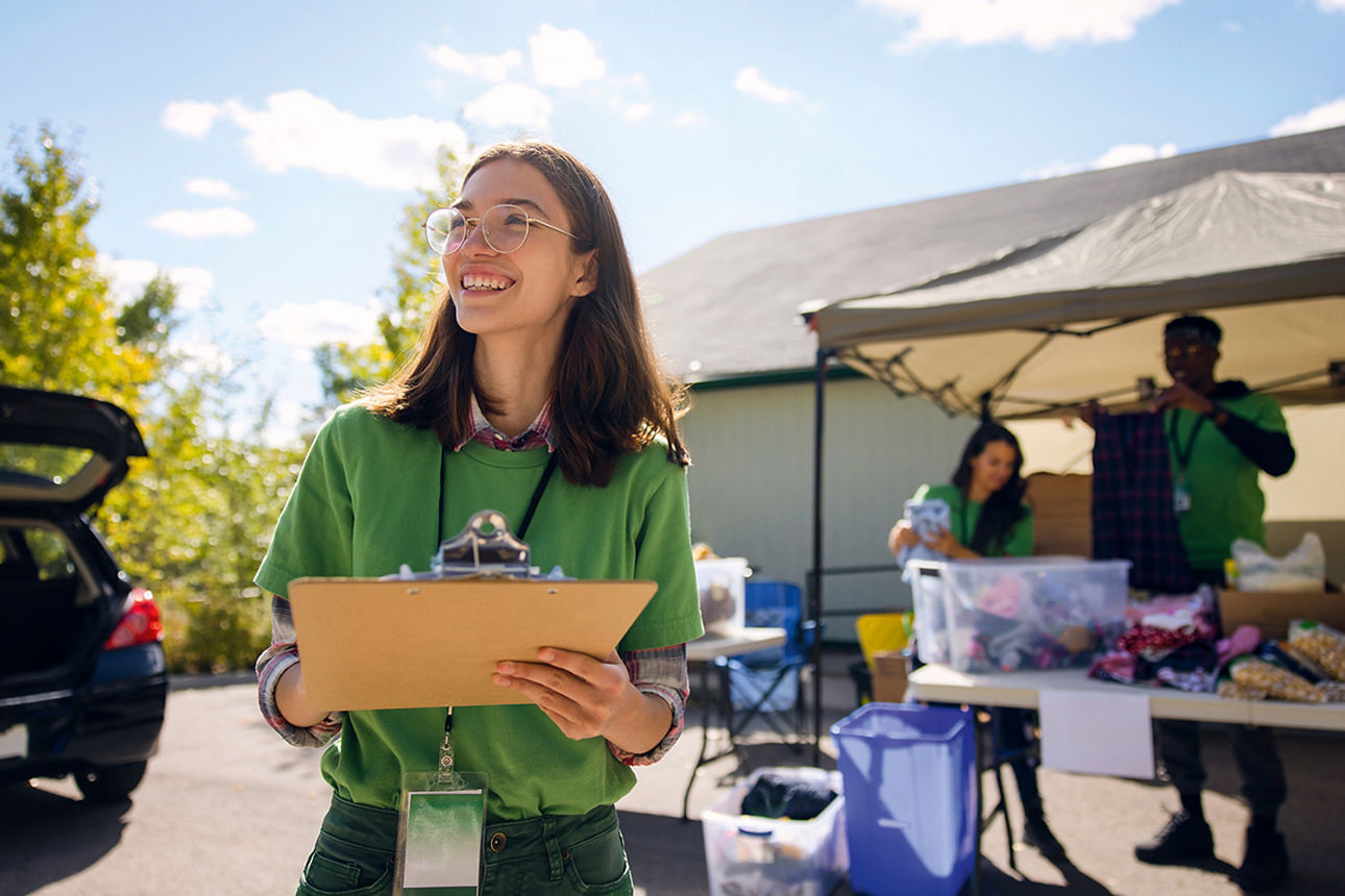 Eine Frau mit einem Schreibbrett und Lanyard steht vor einer Pagode. Unter der Pagode werden Textilien in Kisten gepackt.