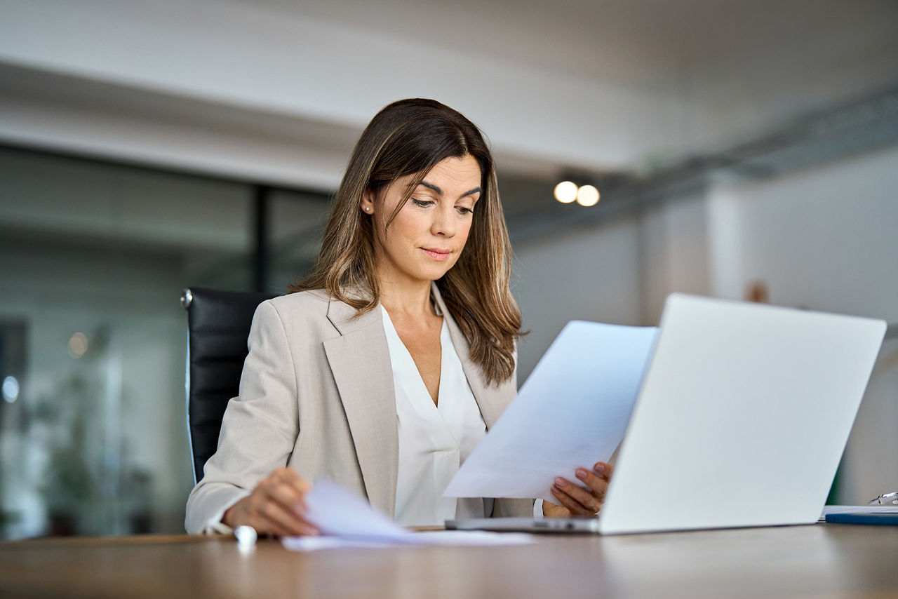 Eine Frau sitzt an einem Schreibtisch und hält Dokumente in der Hand. Vor ihr steht ein Laptop.