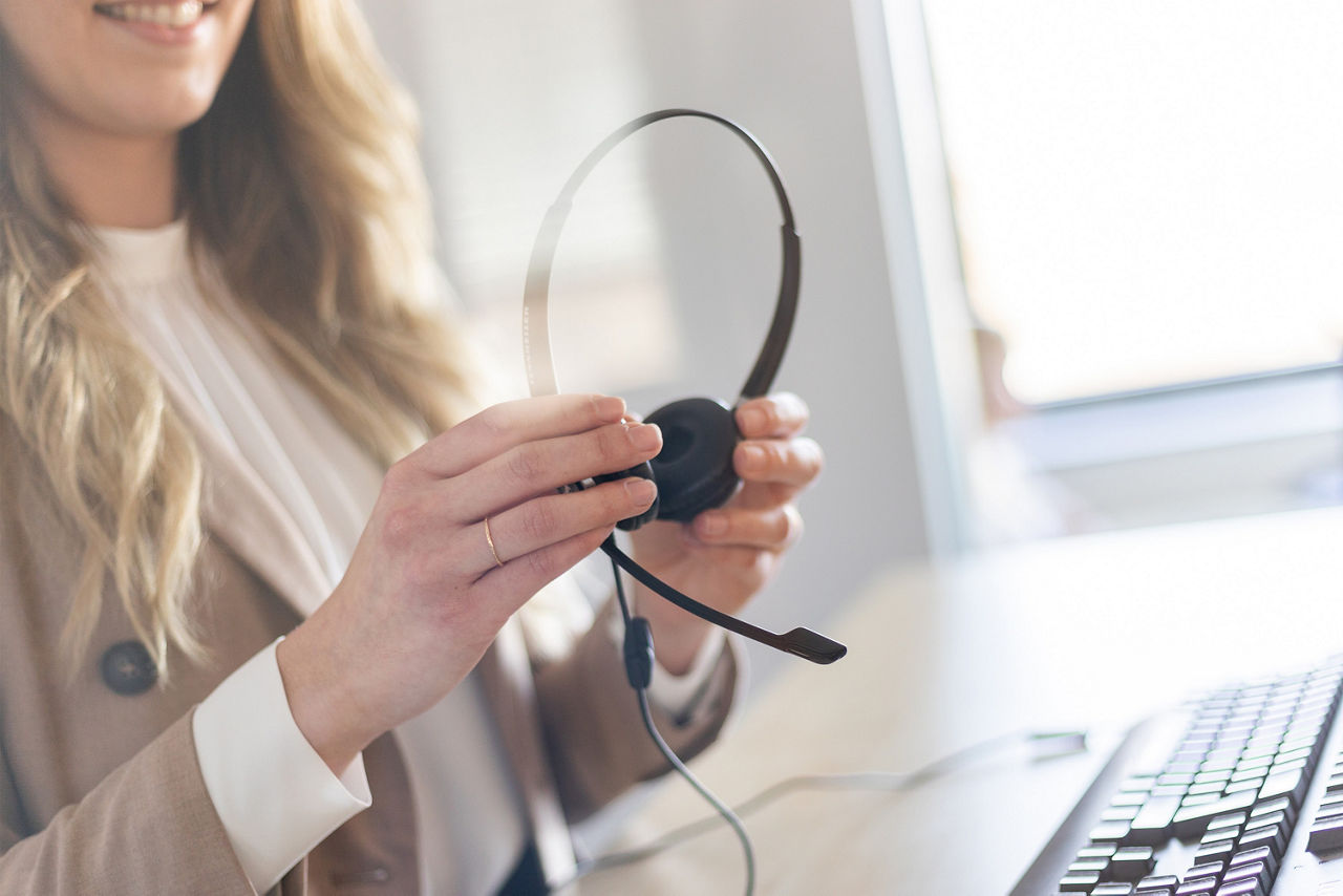 Eine Frau mit Blazer steht vor einem Schreibtisch und hält ein Headset in der Hand.
