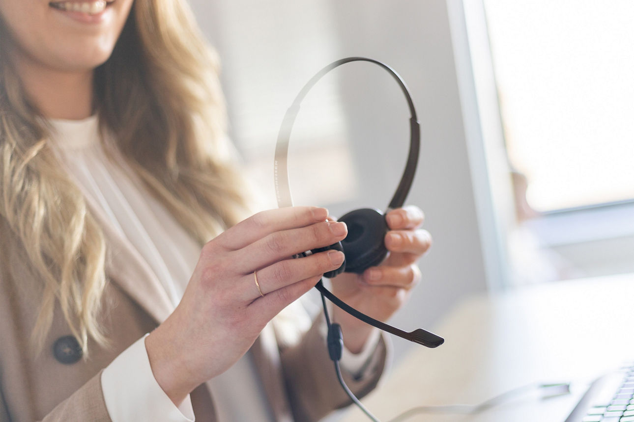 Eine Frau mit Blazer steht vor einem Schreibtisch und hält ein Headset in der Hand.