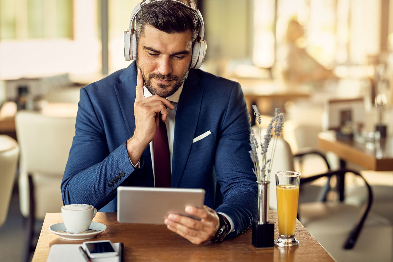 Ein Man in Anzug sitzt an einem Tisch in einem Kaffee und hört mit Over-Ear-Kopfhörern einen Podcast.