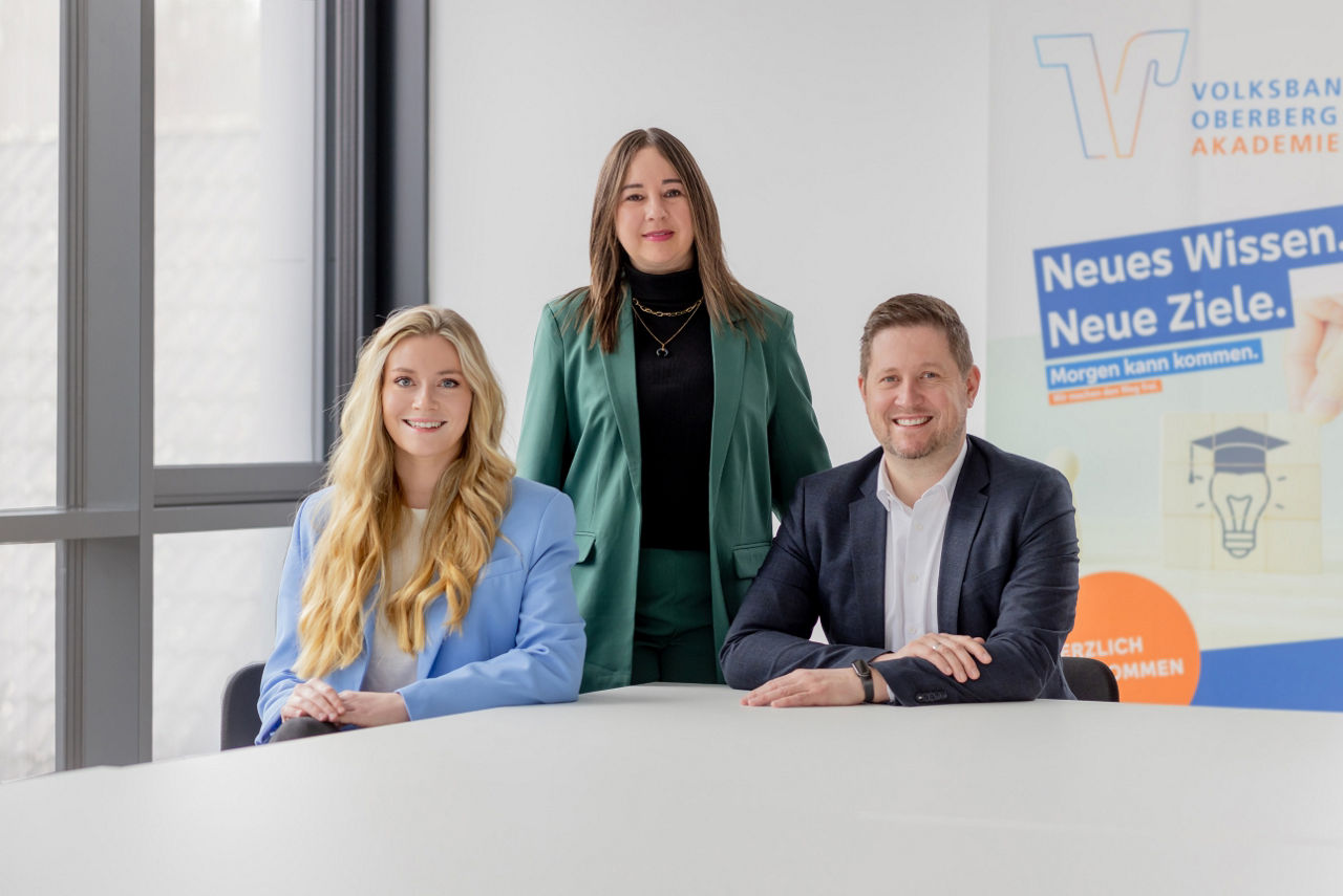 Isabell Stahl steht hinter Laura Harscheid und Mirko Müller, die nebeneinander sitzen. Alle drei blicken freundlich in die Kamera.