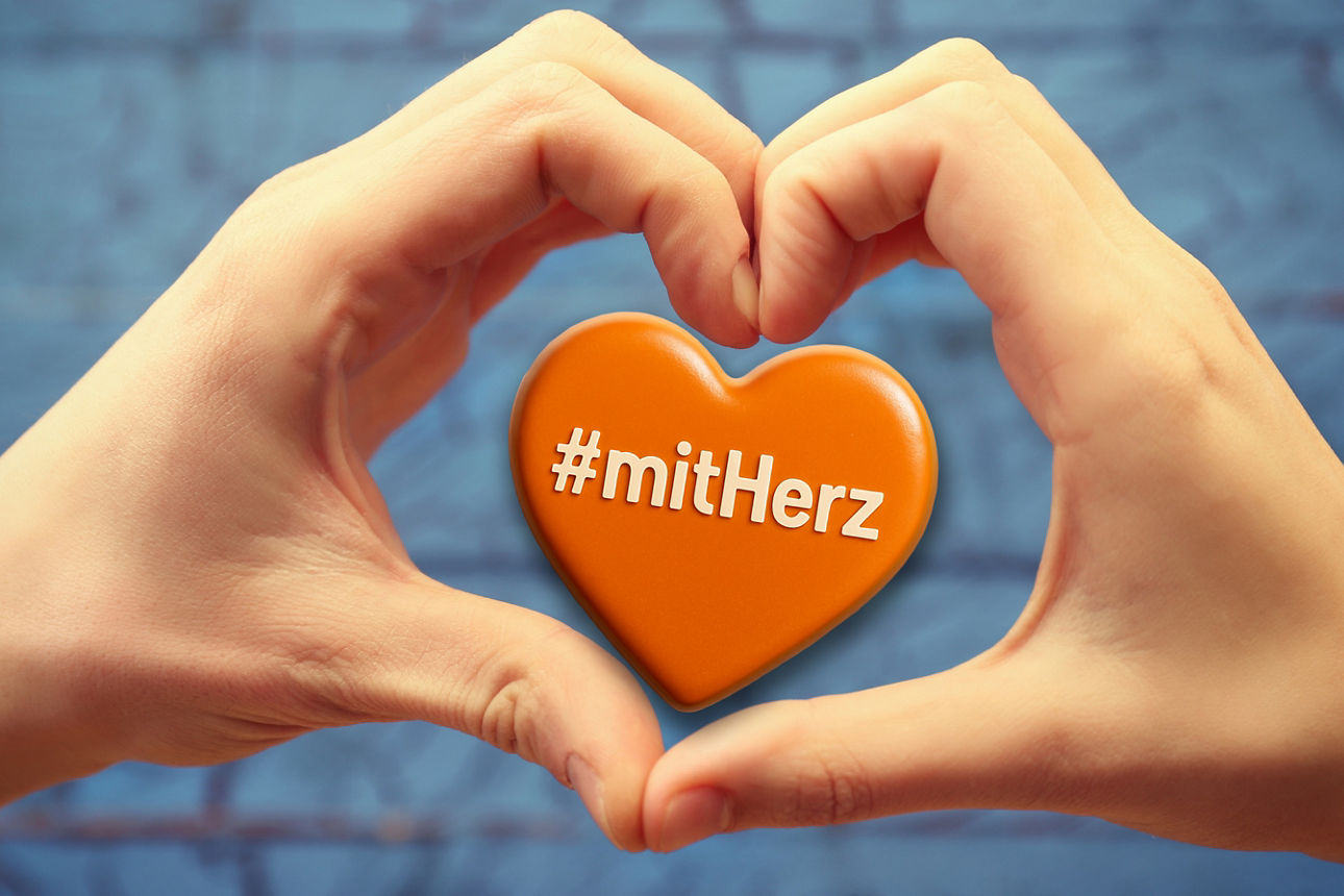 Zwei Hände, die ein Herz formen und in der Mitte ein orangenes Herz mit der Aufschrift "#mitHerz"
