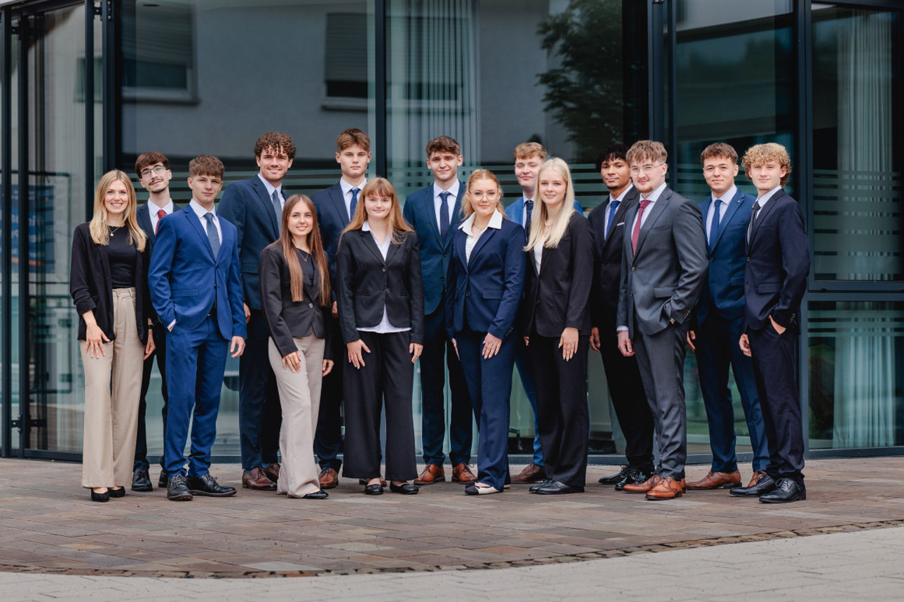 Gruppenfoto vor der Hauptstelle der Volksbank Oberberg in Wiehl. Die 14 neuen Auszubildenden stehen zusammen mit Ausbildungsleiterin Sarina Scherbaum (links) und blicken freundlich in die Kamera.