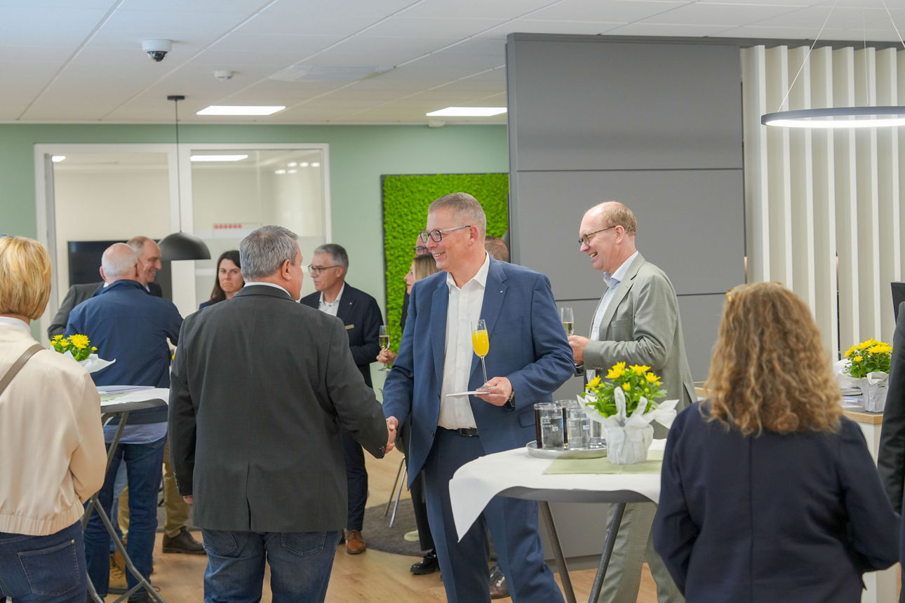 Einige Personen stehen in einem Raum mit Stehtischen, teilweise schütteln sie sich die Hände und lächeln.