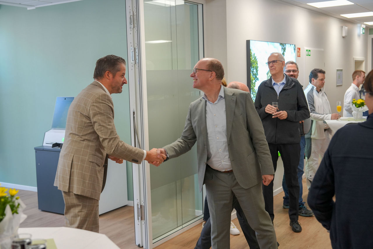 Zwei Männer schütteln sich die Hand und lächeln. Sie stehen in neuen Räumen und im Hintergrund sind weitere Menschen zu sehen