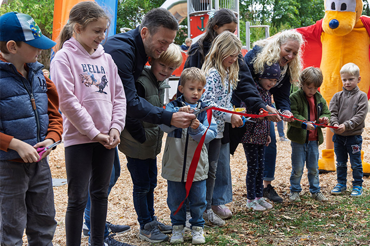 Erwachsene und Kinder auf Spielplatz durchtrennen gemeinsam ein rotes Band