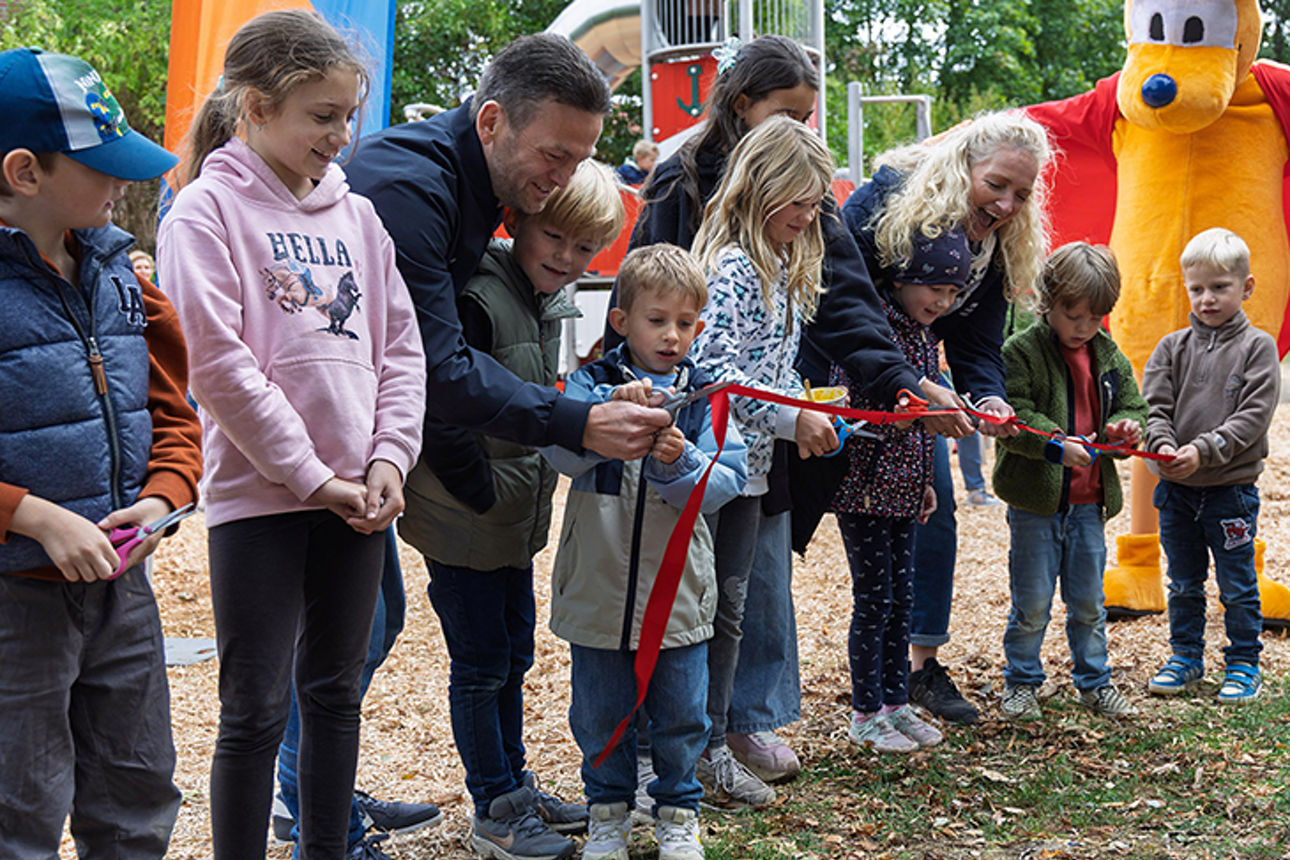 Erwachsene und Kinder auf Spielplatz durchtrennen gemeinsam ein rotes Band