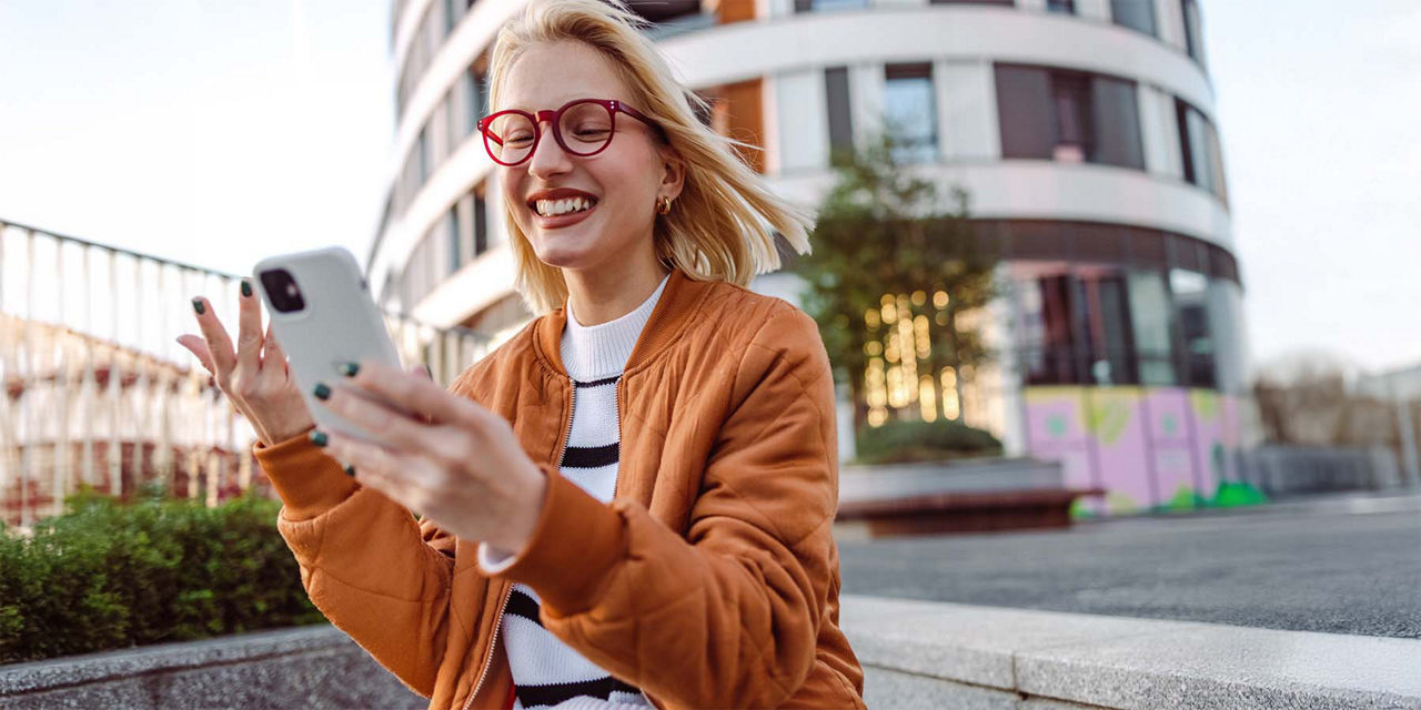 Junge Frau mit roter Brille und blonden Haaren guckt auf Ihr Smartphone und freut sich