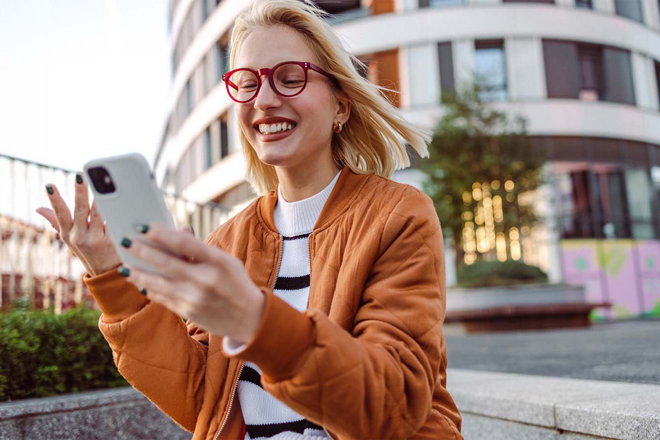  Junge Frau mit roter Brille und blonden Haaren guckt auf Ihr Smartphone und freut sich