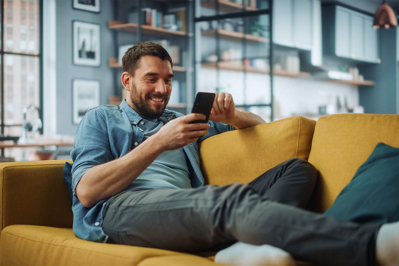 Junger Mann sitzt mit dem Smartphone in der Hand auf dem Sofa