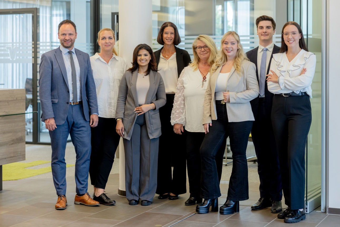 cht Mitarbeitende der Volksbank Oberberg stehen lächelnd in der modernisierten Geschäftsstelle Drabenderhöhe. Sie blicken in die Kamera und freuen sich auf die Wiedereröffnung und den Austausch mit Kundinnen und Kunden.