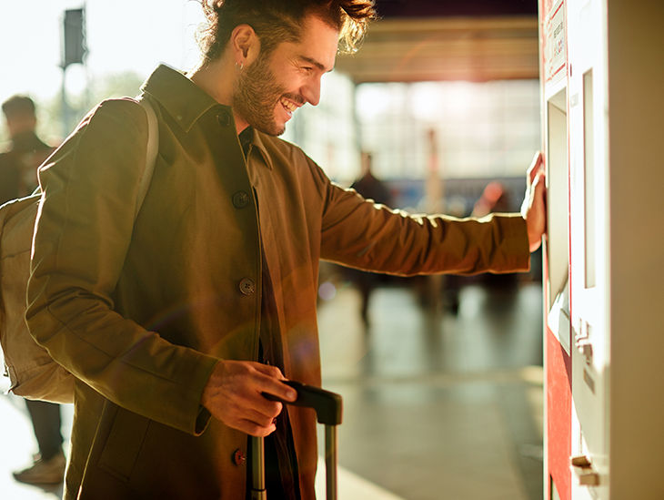 Mann mit Rollkoffer steht an einem Fahrkartenautomaten