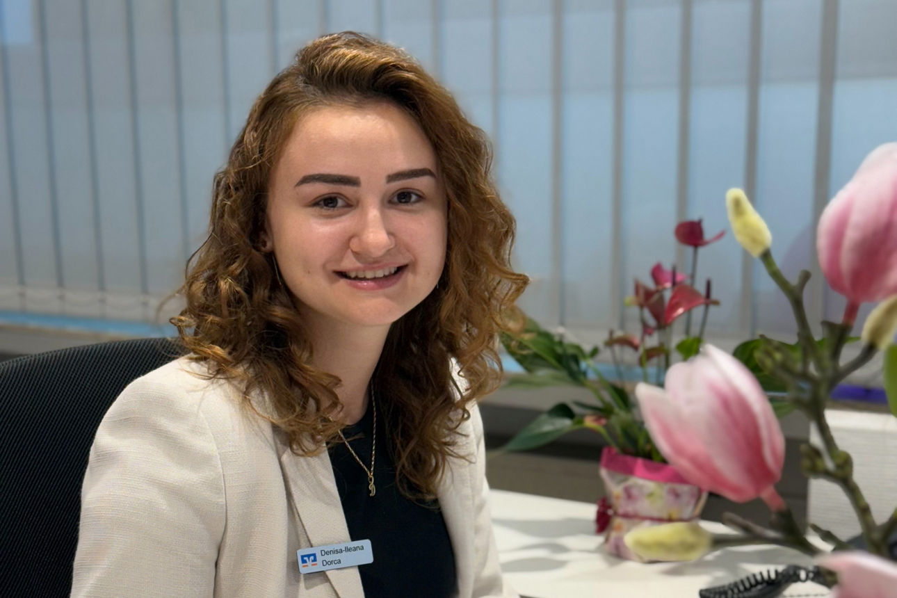 Auszubildende Denisa Dorca sitzt an ihrem Arbeitsplatz in der Geschäftsstelle Bielstein und lächelt in die Kamera.