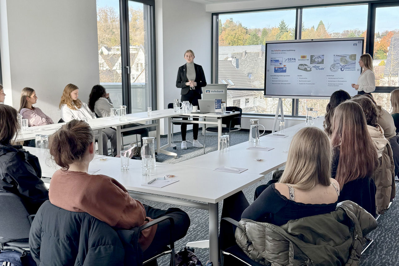 Vortragssituation in der Volksbank Oberberg Akademie: In einem großen Konferenzraum sitzen Schülerinnen und Schüler in U-Form an Tischen, während Sarina Scherbaum und Annabell Strauch vorne stehen und den Jugendlichen das Ausbildungsangebot der Volksbank