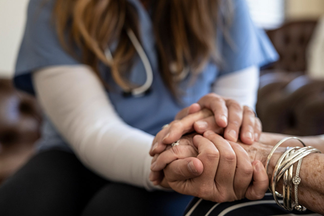 Pflegerin hält Hände einer älteren Frau in wohnlicher Umgebung