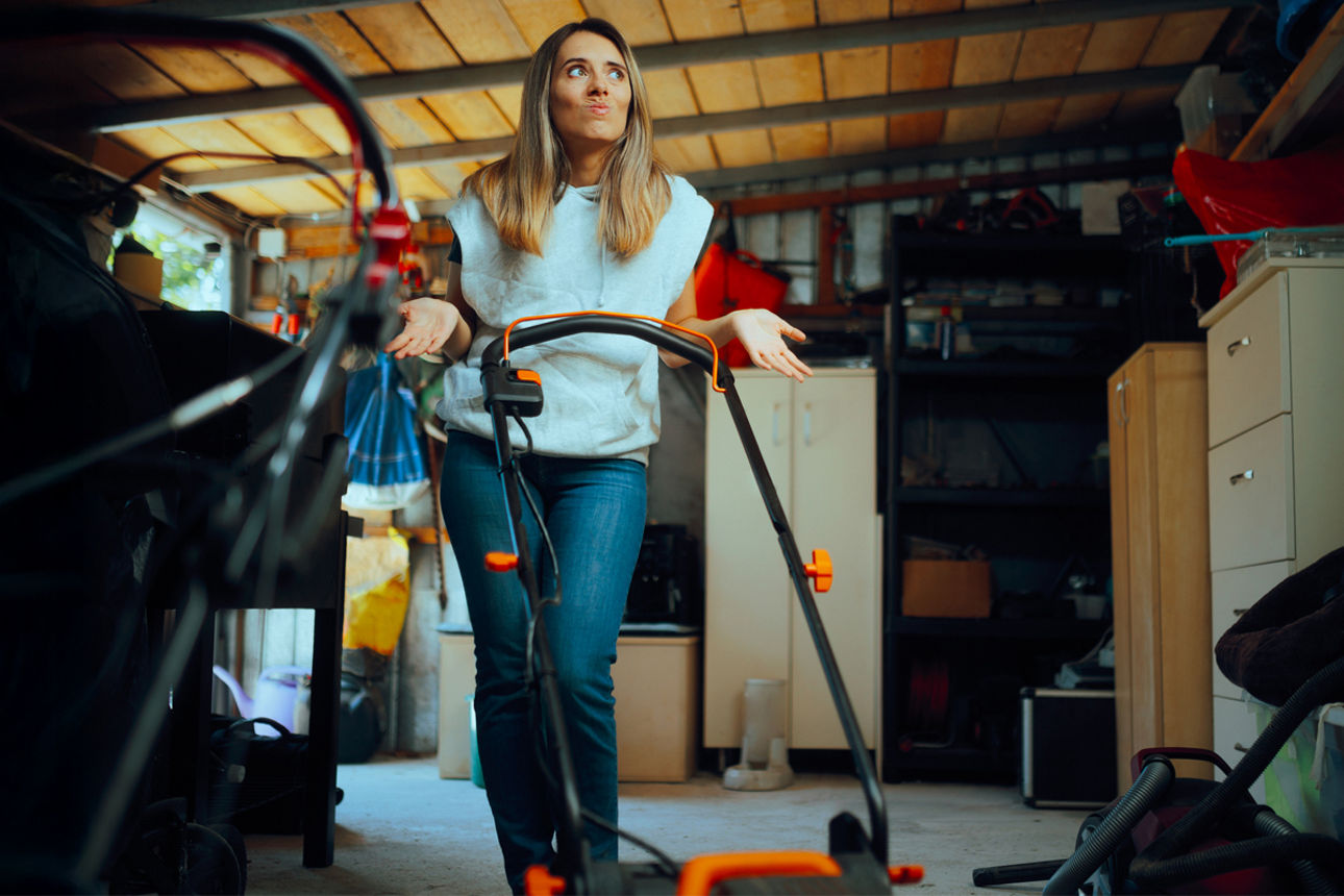 Frau steht fragend mit einem Rasenmäher in einer vollgestellten Garage