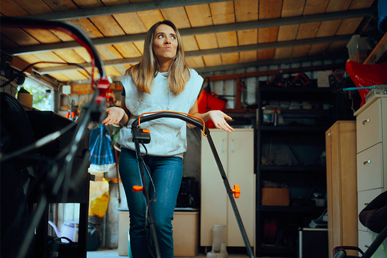 Frau steht fragend mit einem Rasenmäher in einer vollgestellten Garage