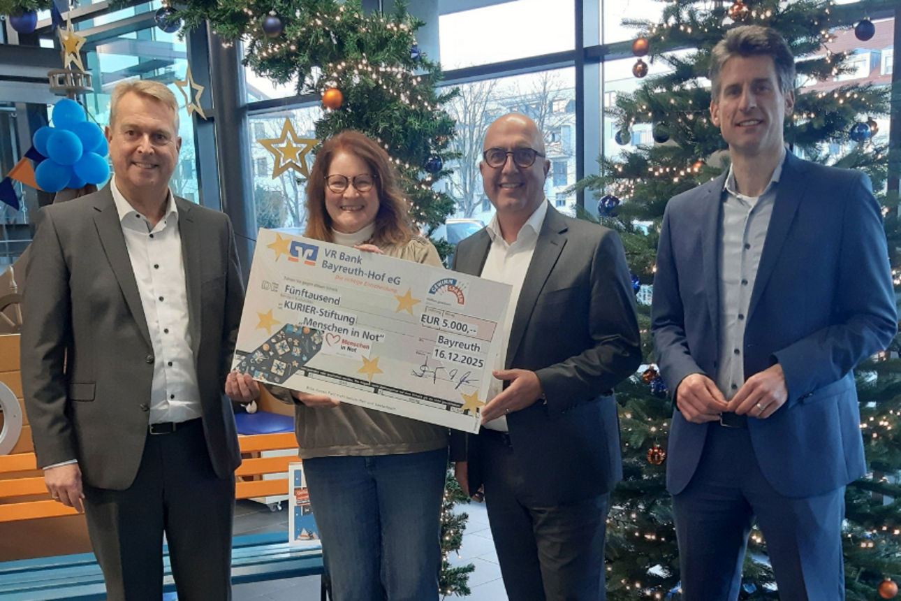 Vier Personen stehen in einem festlich dekorierten Innenbereich mit Weihnachtsbäumen und halten gemeinsam einen Spendenscheck