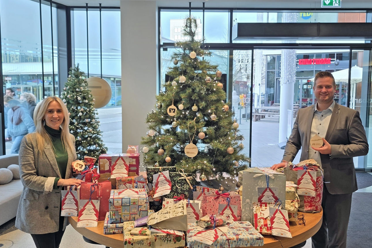 Miriam Elsner und Forum-Geschäftsstellenleiter Justin Wagner stehen im Forum Gummersbach neben einem geschmückten Weihnachtswunschbaum mit vielen verpackten Geschenken aus der Wunschbaumaktion der Volksbank Oberberg.