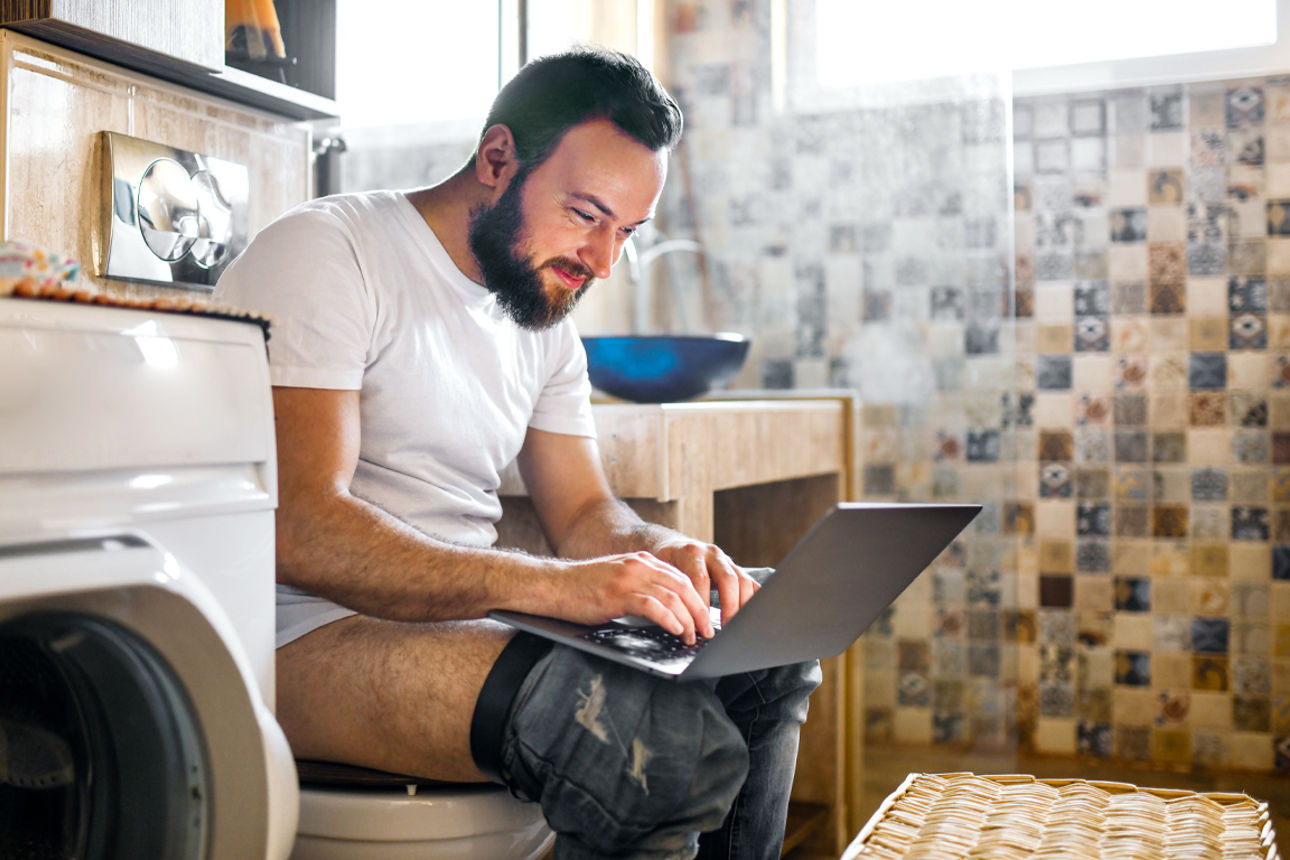 Junger Mann sitzt auf der Toilette und checkt auf dem Handy Aktienkurse.