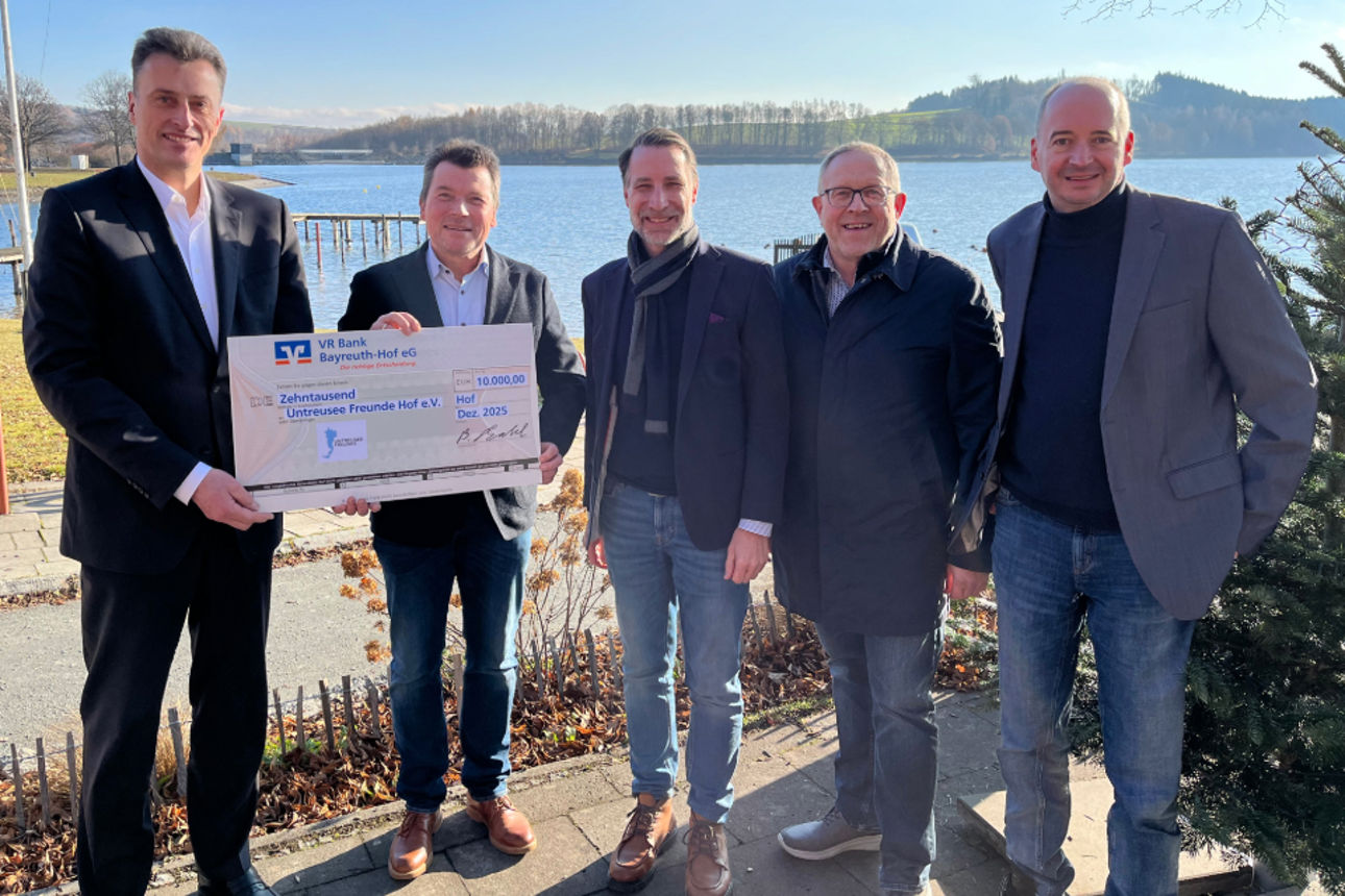 Fünf Personen stehen im Freien vor einem See und halten gemeinsam einen großen Spendenscheck
