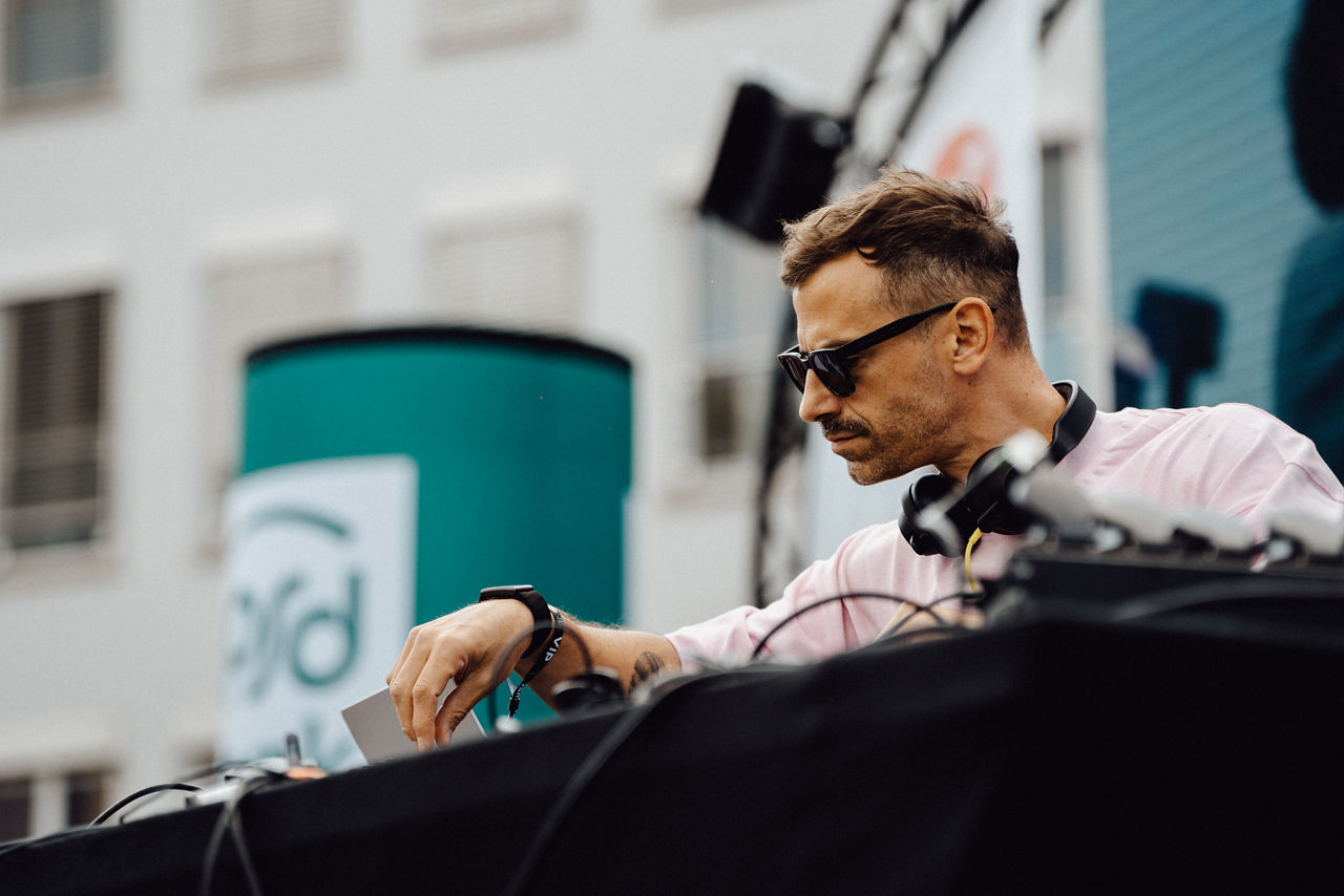 Im Vordergrund ist ein Teil eines DJ-Pults. Ein Mann mixt mit Laptop und Mischpult Musik. Im Hintergrund ist ein Teil der Bühne zu sehen. 